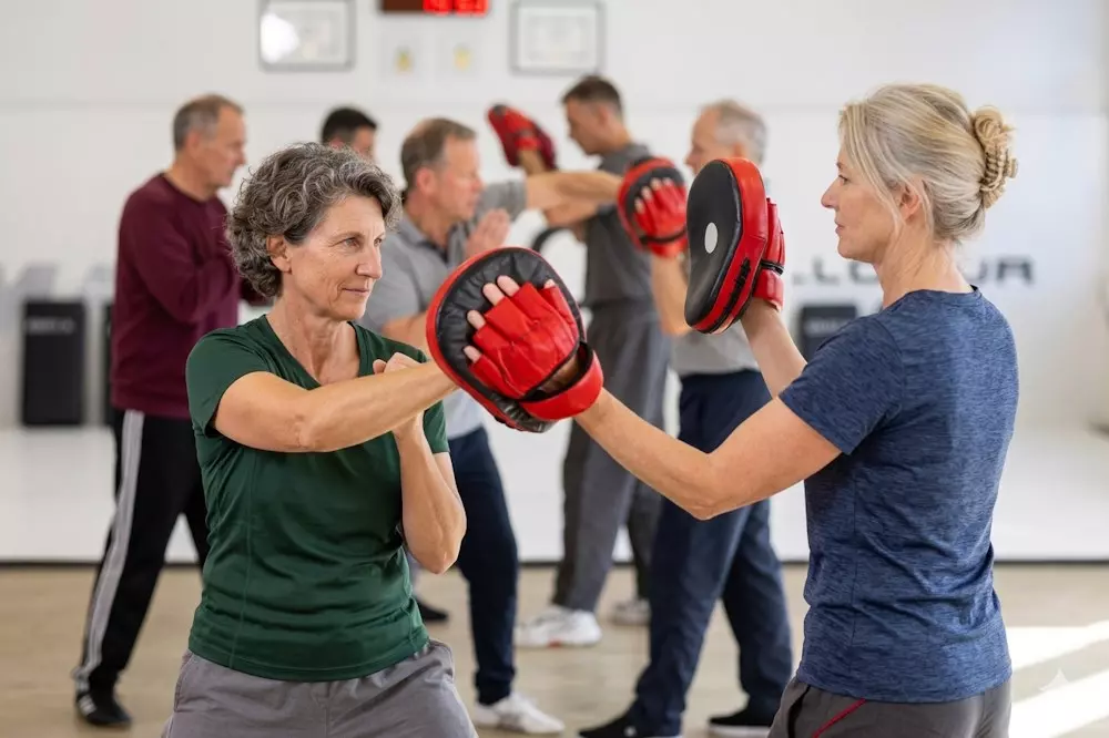 Frauen und Männer ab 50 trainieren gemeinsam Selbstverteidigung mit Schlagpolstern in hellem Trainingsraum